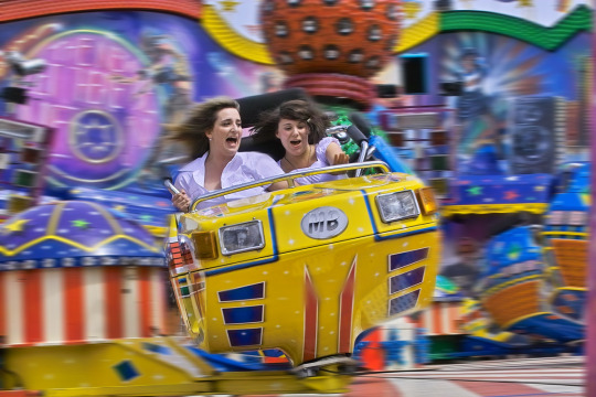 Zwei Besucherinnen werden auf einem modernen Karusell durchgeschüttelt. Mit wehenden Haaren und geöffneten Mündern, sieht man ihnen den Spaß am Nervenkitzel an.