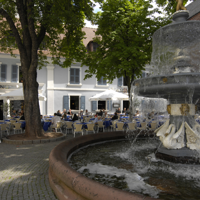 Der Martinsplatz im Sommer. Auf der Außenfläche vom Restaurant St. Martin sitzen bereits einige Leute. Rechts im Bild ist der Martinsbrunnen mit seinen Schwänen zu sehen.