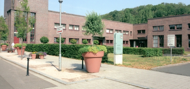 Ein Backsteinbau der Fachhochschule mit geschnittenen Hecken und mehreren bepflanzten kleinen und großen Blumenvasen.