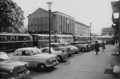 Vor dem Bahnhof stehen dicht gedrängt, hintereinander Busse. Davor stehen parkende Autos. Der heutige Aufbau des Banhofsplatzes ist bereits deutlich zu erkennen.