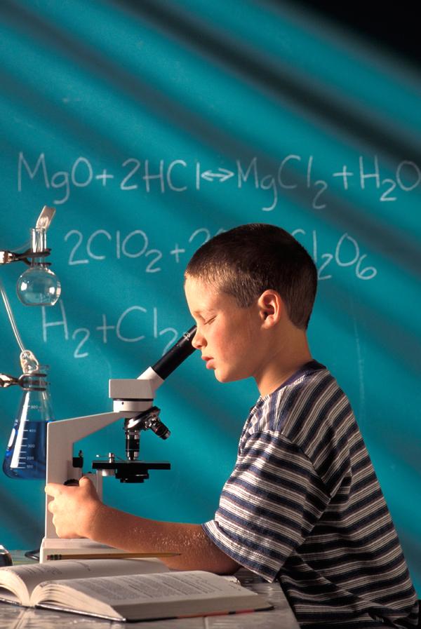 Junge blickt durch ein Mikroskop. An den Seiten des Mikroskops stehen chemische Instrumente,  ein aufgeschlagenes Buch und an der Tafel sind Chemische Verbindungen und Reaktionen notiert