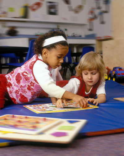 Zwei Mädchen spielen mit einem Zahlenpuzzle. Im Hintergrund sind Stühle, eine Tafel und ein Spielzeugauto zu erkennen.     