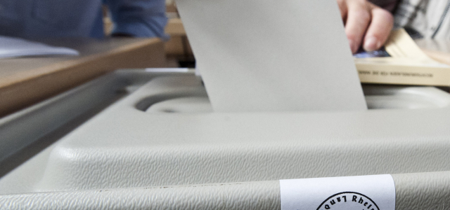 Ein Stimmzettel wird in eine Wahlurne eingeworfen. Das ungebrochene Siegel auf der Urne zeigt, dass die Wahl noch im Gang ist.