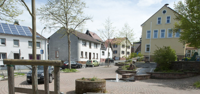 Ein gepflasterter Platz in Dansenberg mit Blick auf die Dansenberger Straße. Auf dem Platz befindet sich ein kleiner, im Boden versunkener, Brunnen und mehrere Bänke als Sitzgelegenheit.