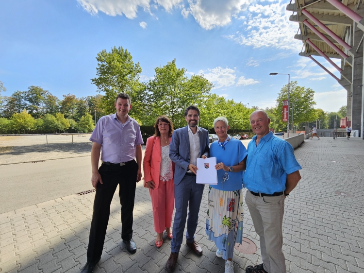Minister Sven Teuber und Oberbürgermeisterin Beate Kimmel trafen sich zur Förderbescheidübergabe vor Ort auf dem Betzenberg. Links Andreas Kühn, Referent im Ministerium für Kommunen, Bauen, Wohnen und Kultur, und Elke Franzreb, Leiterin Referat Stadtentwicklung, rechts Joachim Wilhelm, ebenfalls Referat Stadtentwicklung. Die Gruppe steht vor der Südtribüne des Stadions, im Rücken der Parkplatz Süd, der im Zuge des Verfahrens ebenfalls überplant wird. © Stadt Kaiserslautern