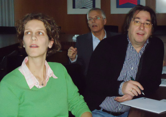 Frau im grünen Pullover und ein Mann im karierten Hemd mit Sakko hören interessiert einem Vortrag zu