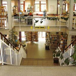 Man steht oben auf der Treppe und kann gleichzeitig den ersten, wie auch zweiten Stock entdecken. In beiden Etagen sind hohe Regale mit Büchern.