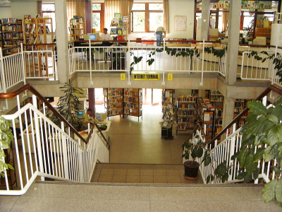 Man steht oben auf der Treppe und kann gleichzeitig den ersten, wie auch zweiten Stock entdecken. In beiden Etagen sind hohe Regale mit Büchern.