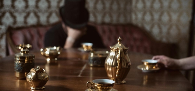 Tisch mit goldenem Teeservice - im Hintergrund ein Mann mit Zylinder, das Gesicht hinter der rechten Hand verbergend.