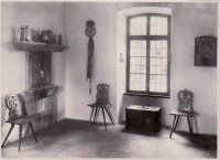 Eine Ecke mit drei kunstvoll geschnitzten Stühlen und einer schweren Truhe. Metallisch glänzendes Geschirr ist als Dekoration auf einem steineren Sims aufgestellt.