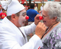 Ein Clown mit roter Nase setzt einer älteren Dame ebenfalls eine rote Nase auf. Diese nimmt es mit viel Humor.