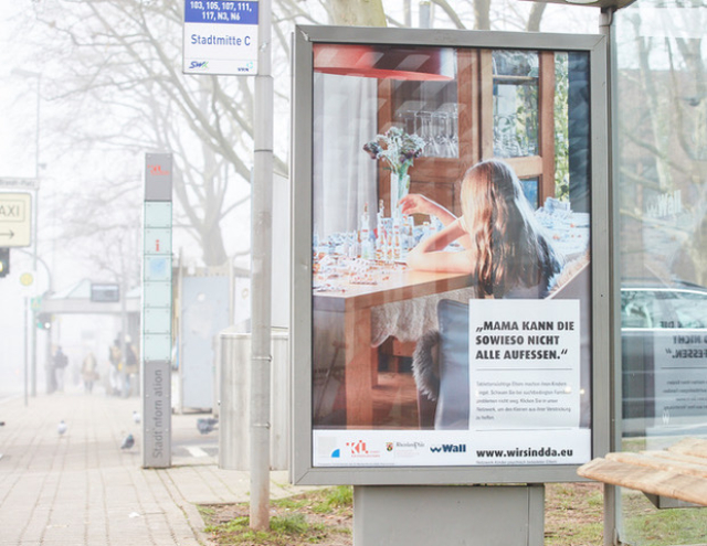 Bushaltestelle mit Plakat. Auf dem Plakat ist ein Kind mit einem Berg von Medikamenten (-schachteln) zu sehen. 