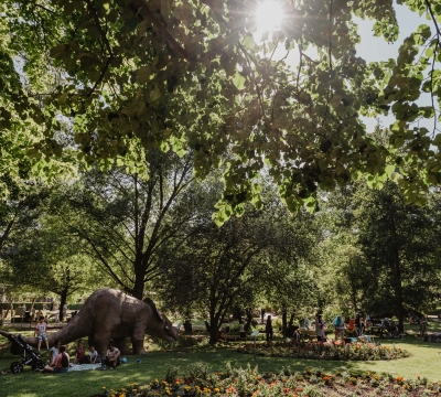 Auf dem Bild sieht man Menschen auf Picknickdecken und einen Dinosaurier. Im Hintergrund viele Bäume. Es ist Sommer und die Sonne scheint.