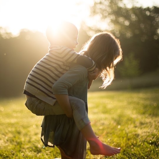 Ein Mädchen trägt einen kleineren Jungen Huckepack. Die Kinder sind auf einer Wiese. Im Hintergrund sieht man Bäume. Die Sonne scheint.