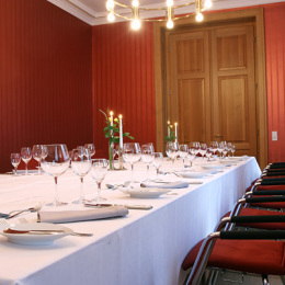 Der Raum besitzt rote Holzvertäfelung an den Wänden und rote Polster auf den Stühlen. Die Tafel ist gedeckt.